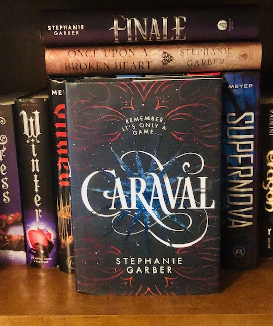 A copy of Caraval sits face forward on a book shelf. On the left are visible spines for books called Cress, Winter, and Gilded. To the right are book spines for Supernova and Eragon. Above Caraval lay the horizontal spines for books titled Finale and Once Upon a Broken Heart. The cover depicts the title written in an elaborate script, with a blue starburst behind it and red line art surrounding everything. The tagline reads: Remeber it's only a game...