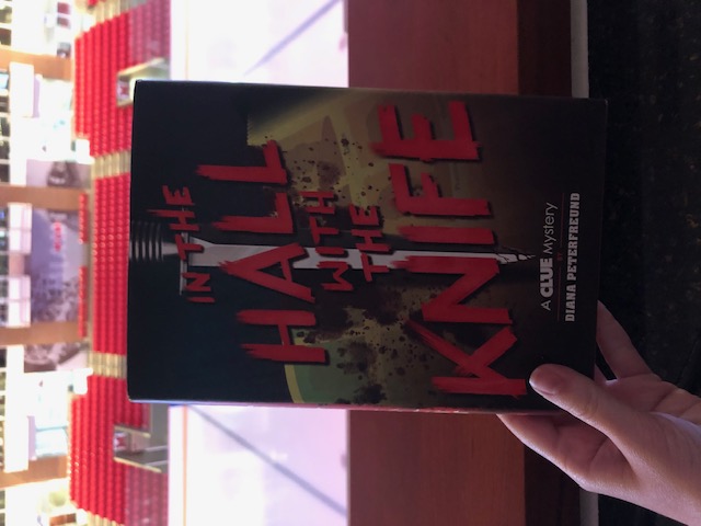 A hand holds In the Hall with Knife in front of the edge of a balcony and a set of stadium seats.