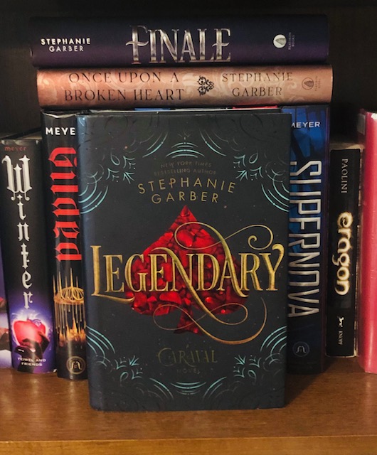 A copy of Legendary sits face forward on a book shelf. On the left are visible spines for books called Winter and Gilded. To the right are book spines for Supernova and Eragon. Above Legendary lay the horizontal spines for books titled Finale and Once Upon a Broken Heart. The cover depicts the title written in an elaborate script, with a red spade behind it and teal line art surrounding everything.