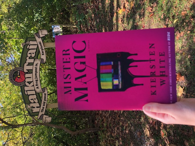 A hand holds a copy of Mister Magic underneath an archway sign that reads: Haiku Trail. Above the title is a crest with a cardinal and beneath it ribbon script that reads: The old pond. A frong jumped in, Kerplunk! A forest lies in the background. The book cover displays a pixelated old-fashioned tv melting at the bottom, on top of a pink background.