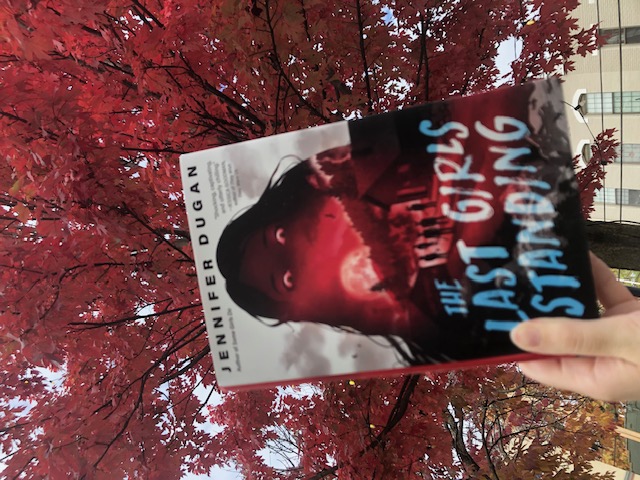 A hand holds a copy of The Last Girls Standing up to a large tree with red leaves.