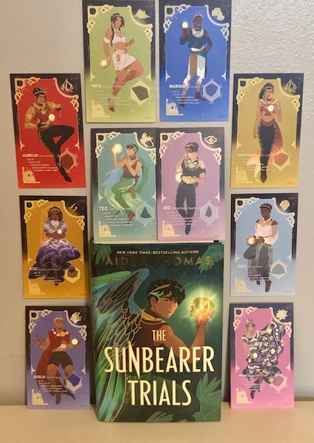 An image of a hardcover copy of the Sunbearer trials leaned agasint a wall surrounded by trading cards of all the contestants. The first row above the books depicts Niya and Marino. The second row contains cards for Teo and Xio. On the left side of the book is Aurelio, Atzi, then Ocelo in a column. On the right side is Auristela, Dezi, and Xochi. The cover depicts Teo looking back over his shoulder, colorful wings displayed, with a sun crown in his hair and a glowing skull in his hand.